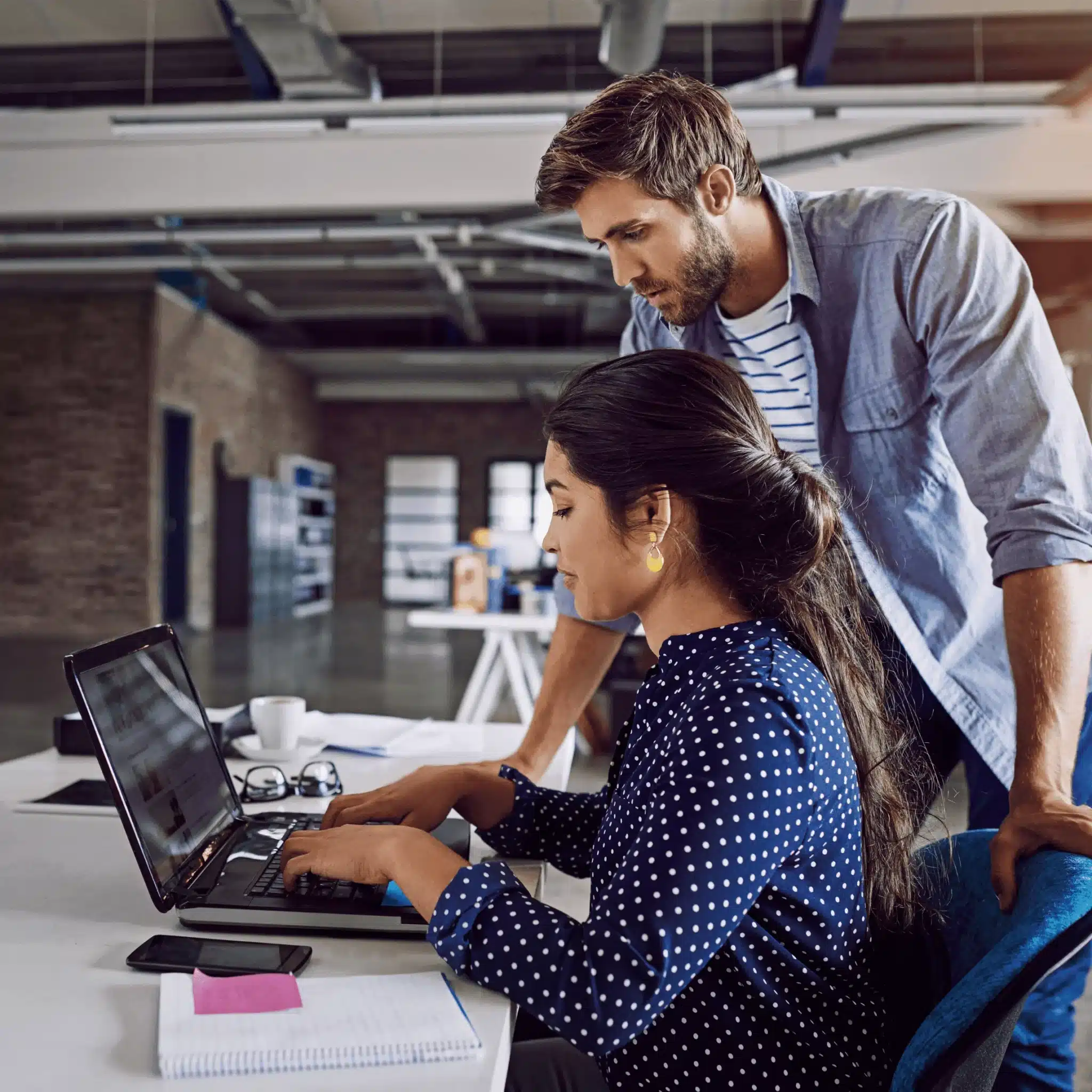 In a modern office setting, a woman types on a laptop at her desk while a man stands beside her, discussing how to empow