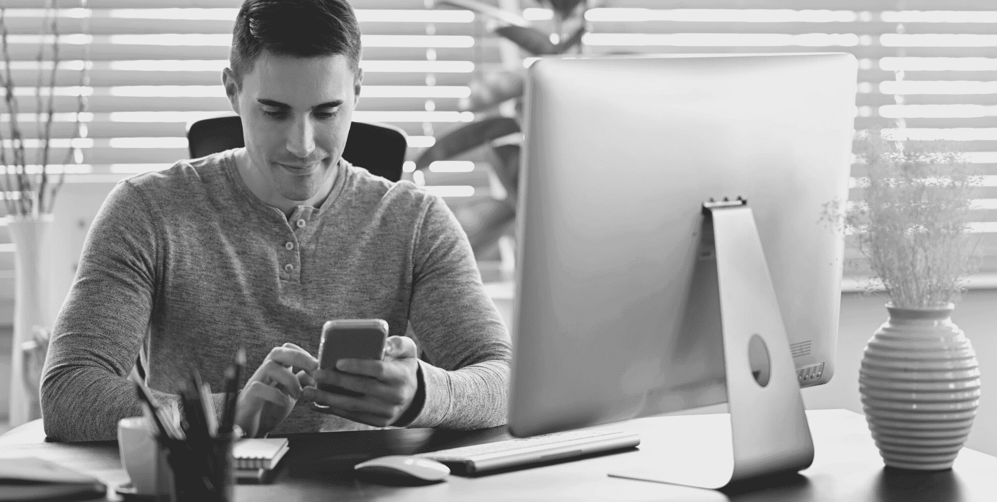 Smart young man using smartphone at modern office desk, working on computer, monochrome.