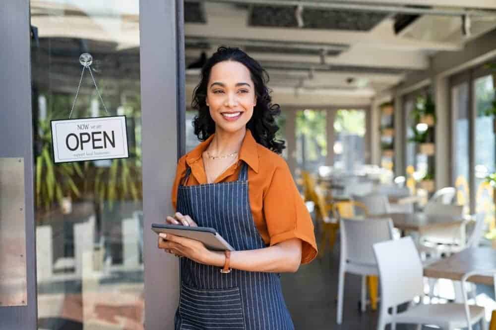 A woman in an apron holding a tablet for digital marketing outside a restaurant.